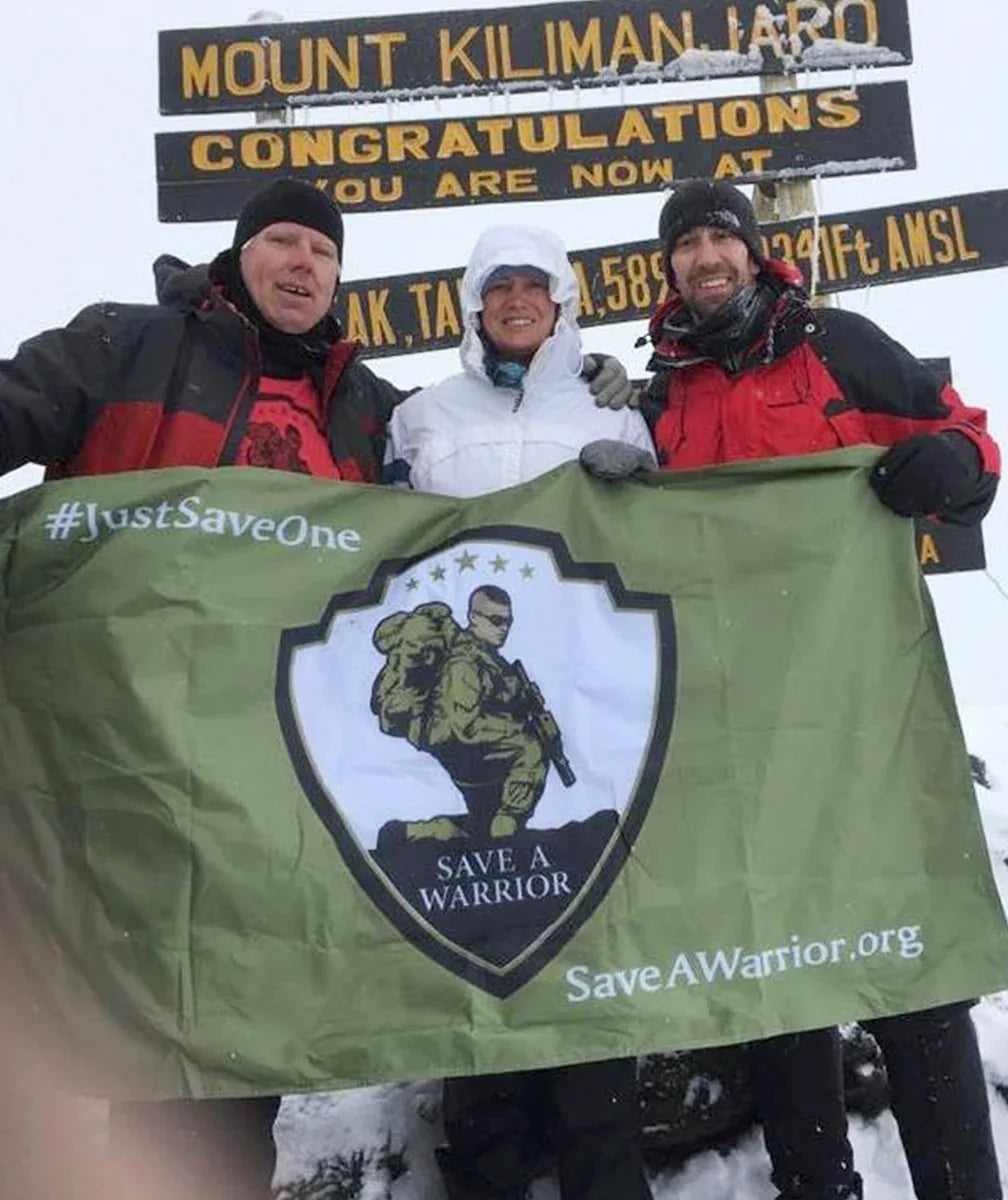 Three people in winter clothing holding a banner with a logo and text in front of a mountain sign.
