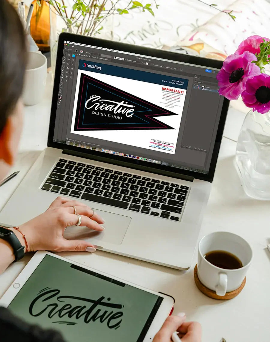 Person working on a laptop with design software, surrounded by a cup of coffee, tablet, and flowers.