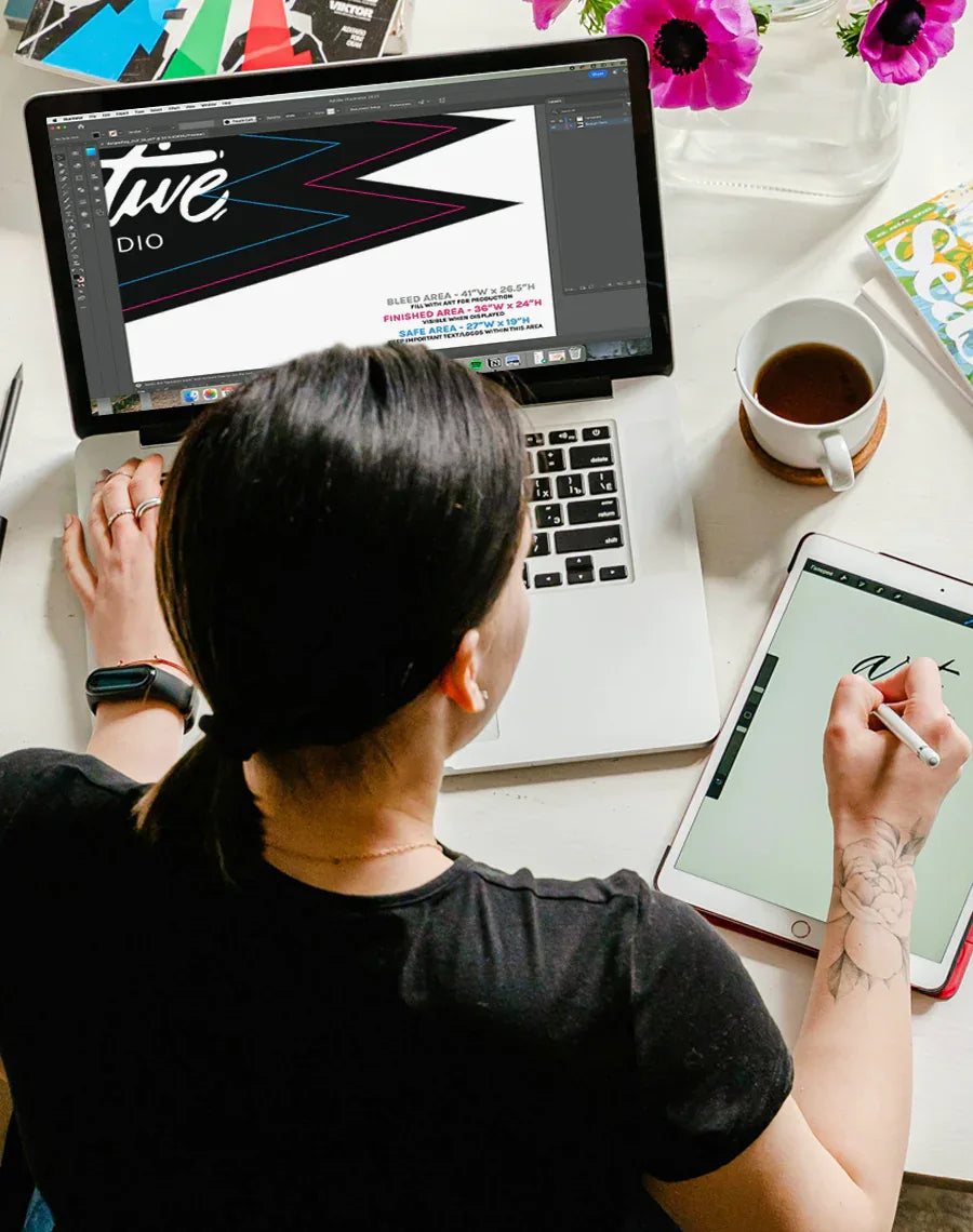 Person using a laptop and tablet with a coffee cup and flowers on a desk