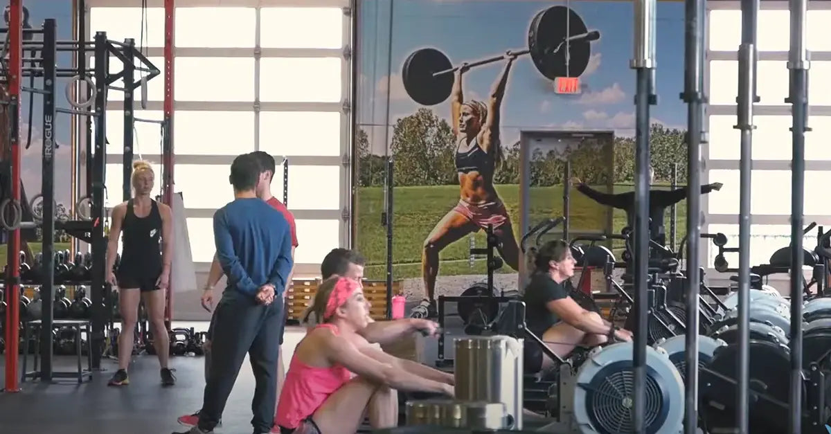 People exercising in a gym with a large wall mural of a woman lifting weights.